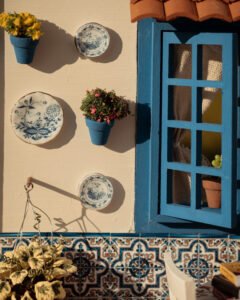 Escena en miniatura detallada de un patio andaluz tradicional que incluye paredes con macetas azules de flores, una ventana azul con tejadillo de barro, azulejos decorativos y una mesa con libros, guitarra y alimentos en miniatura.