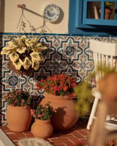 Escena en miniatura detallada de un patio andaluz tradicional que incluye paredes con macetas azules de flores, una ventana azul con tejadillo de barro, azulejos decorativos y una mesa con libros, guitarra y alimentos en miniatura.