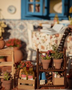 Escena en miniatura detallada de un patio andaluz tradicional que incluye paredes con macetas azules de flores, una ventana azul con tejadillo de barro, azulejos decorativos y una mesa con libros, guitarra y alimentos en miniatura.