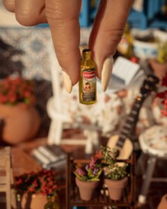 Escena en miniatura detallada de un patio andaluz tradicional que incluye paredes con macetas azules de flores, una ventana azul con tejadillo de barro, azulejos decorativos y una mesa con libros, guitarra y alimentos en miniatura.