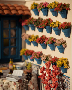 Escena en miniatura detallada de un patio andaluz tradicional que incluye paredes con macetas azules de flores, una ventana azul con tejadillo de barro, azulejos decorativos y una mesa con libros, guitarra y alimentos en miniatura.