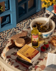 Escena en miniatura detallada de un patio andaluz tradicional que incluye paredes con macetas azules de flores, una ventana azul con tejadillo de barro, azulejos decorativos y una mesa con libros, guitarra y alimentos en miniatura.