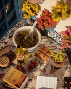 Escena en miniatura detallada de un patio andaluz tradicional que incluye paredes con macetas azules de flores, una ventana azul con tejadillo de barro, azulejos decorativos y una mesa con libros, guitarra y alimentos en miniatura.