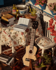 Escena en miniatura detallada de un patio andaluz tradicional que incluye paredes con macetas azules de flores, una ventana azul con tejadillo de barro, azulejos decorativos y una mesa con libros, guitarra y alimentos en miniatura.