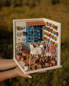 Escena en miniatura detallada de un patio andaluz tradicional que incluye paredes con macetas azules de flores, una ventana azul con tejadillo de barro, azulejos decorativos y una mesa con libros, guitarra y alimentos en miniatura.