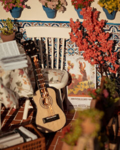 Escena en miniatura detallada de un patio andaluz tradicional que incluye paredes con macetas azules de flores, una ventana azul con tejadillo de barro, azulejos decorativos y una mesa con libros, guitarra y alimentos en miniatura.
