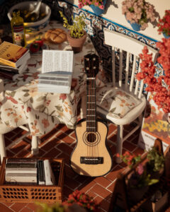 Escena en miniatura detallada de un patio andaluz tradicional que incluye paredes con macetas azules de flores, una ventana azul con tejadillo de barro, azulejos decorativos y una mesa con libros, guitarra y alimentos en miniatura.