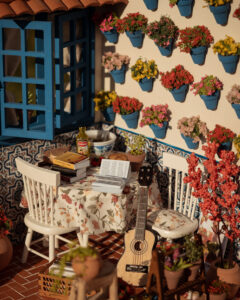 Escena en miniatura detallada de un patio andaluz tradicional que incluye paredes con macetas azules de flores, una ventana azul con tejadillo de barro, azulejos decorativos y una mesa con libros, guitarra y alimentos en miniatura.