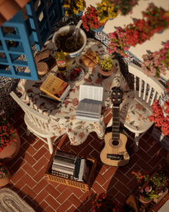 Escena en miniatura detallada de un patio andaluz tradicional que incluye paredes con macetas azules de flores, una ventana azul con tejadillo de barro, azulejos decorativos y una mesa con libros, guitarra y alimentos en miniatura.
