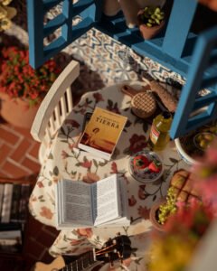 Escena en miniatura detallada de un patio andaluz tradicional que incluye paredes con macetas azules de flores, una ventana azul con tejadillo de barro, azulejos decorativos y una mesa con libros, guitarra y alimentos en miniatura.